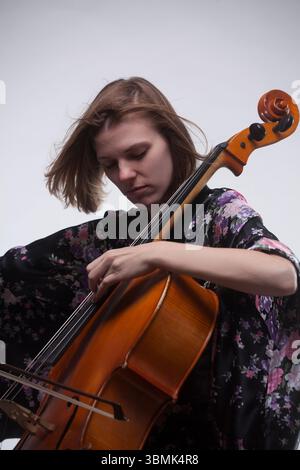 Junge Erwachsene kaukasische Frau spielt Cello im Studio, trägt ein schwarzes Blumengewand, konzentriert. Stockfoto