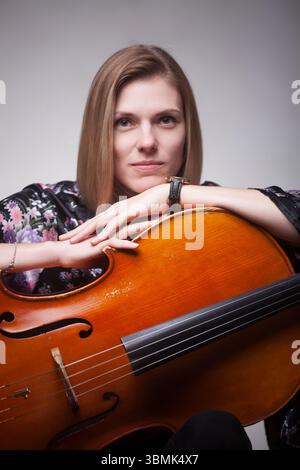 Erwachsene kaukasische Frau in Blumenoberteil, lehnt sich auf ein Cello in einem Studio-Porträt und blickt in die Kamera. Stockfoto