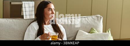 Eine junge, schöne Frau sitzt bequem zu Hause und genießt ein warmes Getränk, während sie sich entspannt. Stockfoto