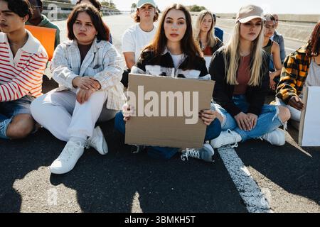 Eine Gruppe junger Erwachsener, die zusammen auf einer Straße mit Protestschildern sitzen und sich für eine Sache bei hellem Sonnenlicht einsetzen. Stockfoto