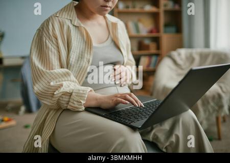 Schwangere asiatische Frau, die drinnen sitzt, mit einem Laptop mit einer Hand, während sie sanft den Bauch berührt, sich auf den Bildschirm konzentriert, lässiges Zuhause, Vorbereitung auf die Mutterschaft, junger Erwachsener Stockfoto