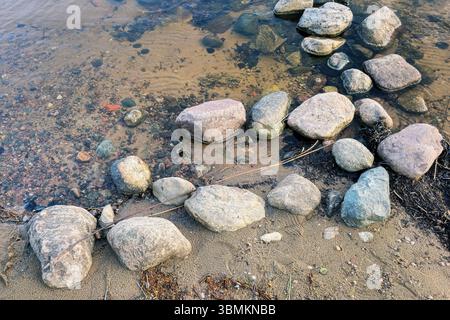 Große und kleine Steine liegen an einem Sommertag auf dem Sand und in klarem Wasser am Ufer eines Sees. Stockfoto