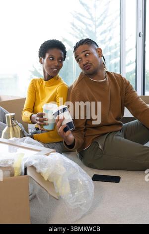 Verschiedene Paare, die Umzugskartons auf Teppich für Fenster auspacken, mit wiederverwendbaren Reisebechern Stockfoto