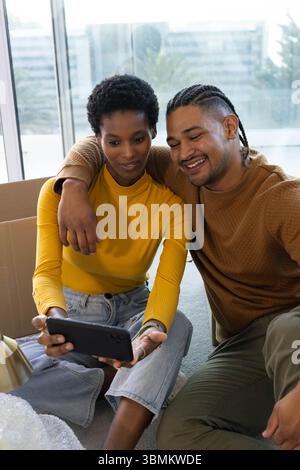 Ein vielseitiges Paar, das Smartphone auf dem Teppichboden neben dem Fenster ansieht, mit Umzugskartons Stockfoto