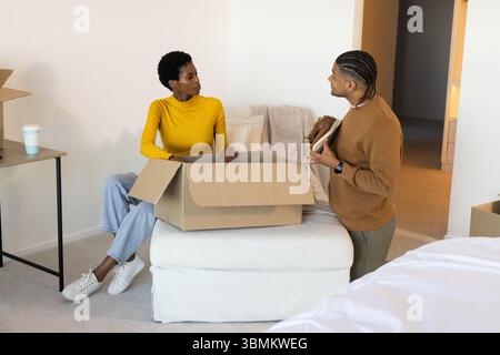 Verschiedene Paare packen Pappkartons im Schlafzimmer aus, halten Schuhe und Kaffeetasse auf Ottomane Stockfoto