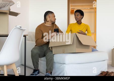 Auspacken diverser Paare, die im Wohnzimmer auf einer weißen Ottomane sitzen, mit Pappkarton und Smartphone Stockfoto
