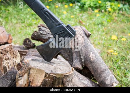 Outdoor-Szene mit einer Axt, die in einem Häckselblock neben einem Holzhaufen eingebettet ist, was auf rustikales Wohnen, Brennholzzubereitung oder Waldarbeiten hindeutet. Stockfoto