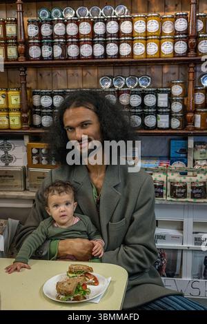 Junger Vater hält Baby im Café und isst ein Sandwich Stockfoto