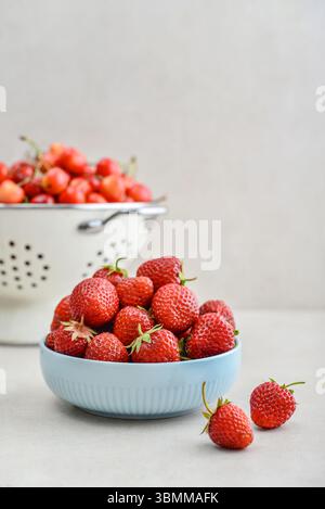 Reife Erdbeeren in einer blauen Schüssel und ein weißes Emailsieb voller süßer Kirschen auf hellem Hintergrund Stockfoto