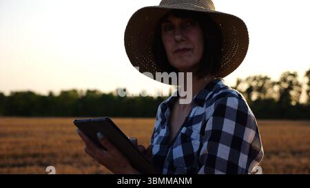 Agrostomistin im Strohhut, die die Ernte auf dem Gerstenfeld bei Sonnenuntergang überwacht. Erwachsener Landwirt mit digitaler Tablette auf Weizenwiese in der Abenddämmerung. Schön Stockfoto