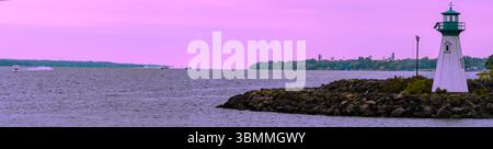 Kleiner weißer und grüner Leuchtturm an einer felsigen Küste unter einem rosa Himmel mit Vergnügungsbooten auf dem St. Lawrence River. Stockfoto