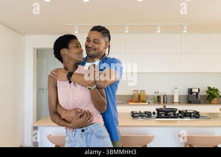 Das vielfältige Paar umarmt die Arme um den Partner auf der Insel in einer modernen Küche mit Herd Stockfoto