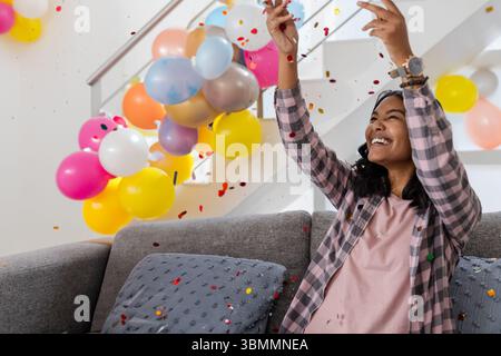 Frau, die auf Couch mit Kissen zwischen bunten Ballons und Konfetti im Wohnzimmer sitzt, Kopierraum Stockfoto