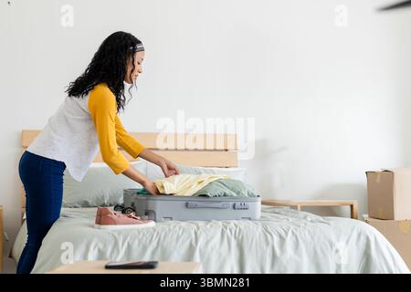 Die Person packt Kleidung in einen offenen Koffer auf einem Holzbett mit pinkfarbenen Turnschuhen im aufgeräumten Schlafzimmer Stockfoto