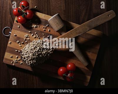 Ein rustikales Stillleben mit rohen Weizenkörnern in Holzlöffeln, frischen Kirschtomaten und Vintage-Küchenutensilien. Stockfoto