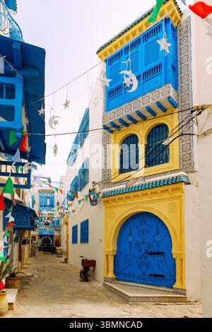 Gasse mit blau verzierten Holztüren, arabischen Balkonen und Fenstern und farbenfrohen Fahnen in der Medina von Hammamet, Tunesien Stockfoto