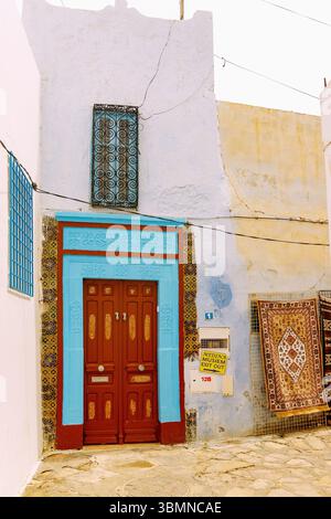Traditionelles Haus mit farbenfroher Holztür mit arabischen Schnitzereien und gewebten Teppichen zum Verkauf an der Hauswand in Hammamet, Tunesien Stockfoto