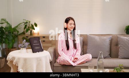 Junge asiatische Frau meditiert zu Hause mit Kopfhörern und hört entspannende Naturgeräusche auf einem Tablet, die Wohlbefinden und Stressabbau fördern Stockfoto