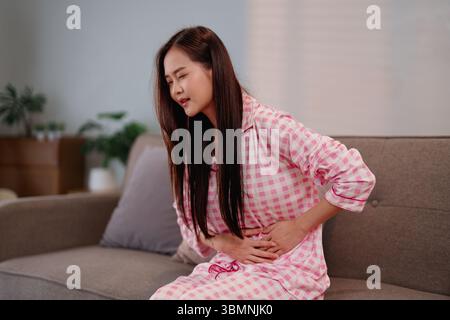 Weibliche Erwachsene mit akuten Magenschmerzen, während sie sich auf der Couch in ihrem Haus ausruhen und sofortige Linderung für gesundheitliche Probleme suchen Stockfoto