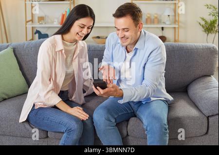 Fröhliches junges Paar, das zu Hause auf dem Sofa sitzt und Apps durchsucht. Stockfoto