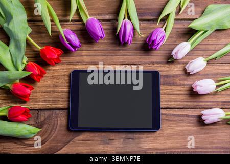 Die Tablette sitzt flach auf einer Holzplatte, umgeben von roten violetten und blassrosa Tulpen Stockfoto