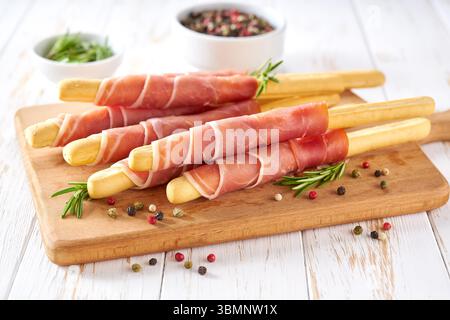 italienisches Stockbrot grissini mit parmaschinken und Rosmarin, traditionelle Brotstangen mit Schinken auf einem weißen Holztisch. Stockfoto