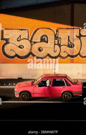 Das leuchtend pinkfarbene Auto fährt vor einer auffälligen Graffiti-Wand mit auffälligen Schriftzügen vorbei Stockfoto