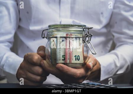Der Mann übergibt Auslagen, Banknoten von Euro-Bargeld aus dem Glasgefäß in der Sparkasse auf dem Rechner. Nahaufnahme von Händen, unerkennbarer Geschäftsmann. Speichern Stockfoto
