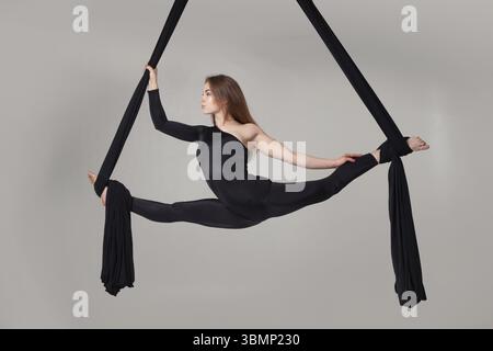Turnerleistung auf schwarzen Leinwänden. Die entzückende Frau in einem dunklen Sportanzug hängt an einem Seil in einem Studio isoliert auf weißem Hintergrund Stockfoto