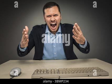 Porträt eines wütenden, verärgerten jungen Mannes in blauem Hemd und Jacke mit erhobenen Händen, der an einem Schreibtisch in der Nähe eines Computers sitzt, isoliert auf grauem Hintergrund. Negati Stockfoto