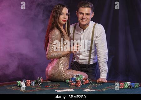 Eleganter Mann und Mädchen spielen Poker im Casino, halten Champagner, feiern ihren Sieg am Tisch mit Stapeln Chips und Karten darauf. Schwarz, rauchig Stockfoto