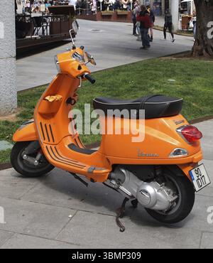 Ein oranger Vespa-Roller parkt auf einem Gehweg in der Nähe eines grasbewachsenen Gebietes, mit verschwommenen Menschen im Hintergrund Stockfoto