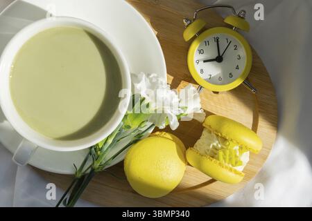 Gelber Wecker Weckzeit Weckruf Süße gelbe Macaron Dessert Tasse grüner Matcha Tee Latte auf Pflanzenbasis in gemütlicher Morgenliegenatmosphäre. Kreativer Urlaub Stockfoto
