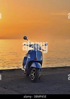 Blauer Vespa-Roller parkte am Strand bei einem pulsierenden Sonnenuntergang, der ein Gefühl von Freiheit und Abenteuer weckt Stockfoto