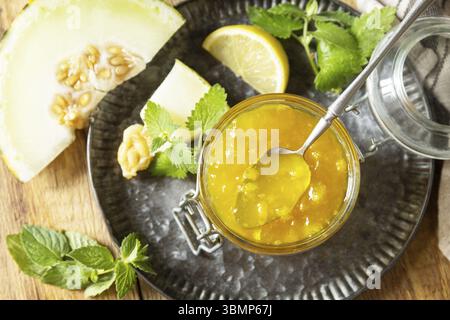 Hausgemachtes Preserve. Süße Melonen- und Zitrusmarmelade oder Gelee in einem kleinen Glas mit frischen Melonenscheiben auf rustikalem Holztisch. Blick von oben Stockfoto