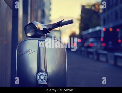 Klassischer Vespa-Roller, der auf einer Stadtstraße mit verschwommenem Verkehr im Hintergrund bei Sonnenuntergang geparkt ist Stockfoto