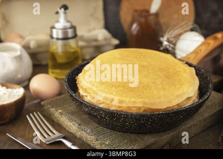 Pancake Day feiern, gesundes Frühstück kochen. Köstliche hausgemachte Crepes oder Pfannkuchen in einer Pfanne und Zutaten auf einem rustikalen Tisch Stockfoto