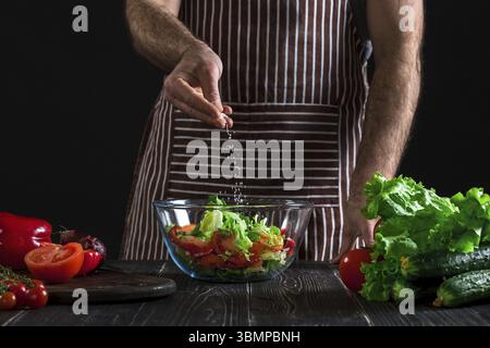 Mann, der gesunden Salat von frischem Gemüse auf einem Holztisch zubereitet. Auf schwarzem Hintergrund. Vegetarisches Essen, gesund oder Kochkonzept Stockfoto