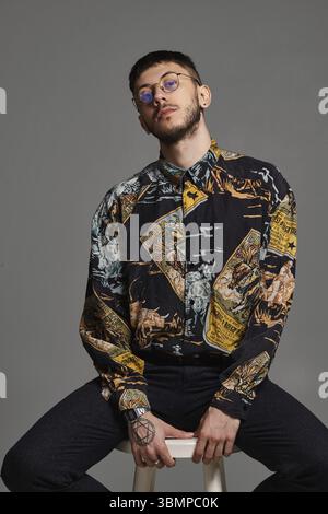 Nahporträt eines hübschen Brünetten in Brille, getragen in einem bunten Hemd, blickt in die Kamera, während er auf einem Barstuhl im Studio sitzt Stockfoto