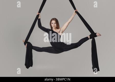 Turnerleistung auf schwarzen Leinwänden. Die entzückende Frau in einem dunklen Sportanzug schaut in die Kamera und hängt in einem Studio an einem Seil Stockfoto