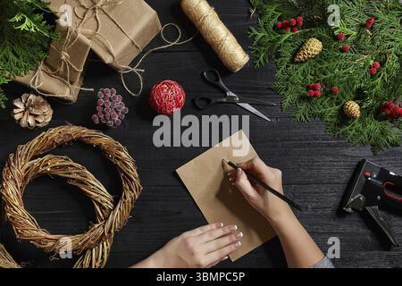 Weihnachten Hintergrund. Draufsicht auf weibliche Hände wickeln Silvester Geschenk. Verpackte Geschenke und Schriftrollen, Fichtenzweige und Werkzeuge auf Holztisch. Arbeitsplatz f Stockfoto
