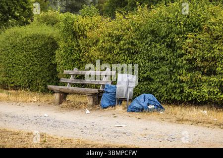 Müll und Müll in Plastiktüten neben einer Parkbank mit Mülleimer auf einer unbefestigten Straße in der Natur Stockfoto