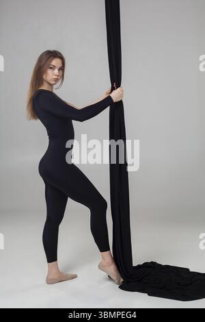 Turnerleistung auf schwarzen Leinwänden. Eine nette Frau in einem dunklen Sportanzug macht akrobatische Elemente an einem Seil und sieht in einem Studi die Kamera an Stockfoto