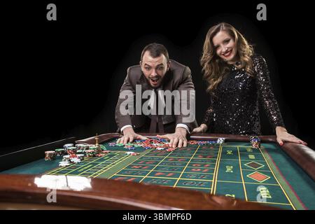 Ein Mann in einem braunen Anzug mit einer schönen Frau in einem schwarzen Kleid spielt an einem Roulette-Tisch in einem Casino. Emotionen von Glücksspielern Stockfoto