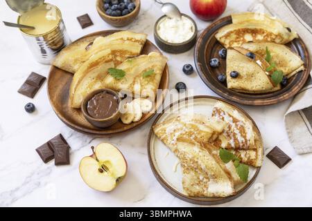Pancake Day feiern, gesundes Frühstück. Hausgemachte süße Crepes mit Heidelbeeren, Ricotta, Äpfeln, Bananen und Schokolade auf einer Marmorplatte Stockfoto