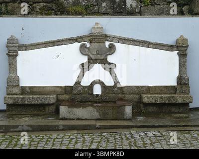 Traditioneller Granitbrunnen mit weiß getünchter Wand, kunstvoll geschnitzten Schnitzereien und einem Kopfsteinpflaster in historischer Umgebung Stockfoto