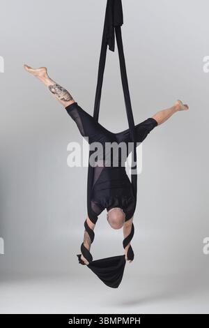 Turnerleistung auf schwarzen Leinwänden. Ein hübscher Glatzkopf in einem dunklen Sportanzug macht akrobatische Elemente, die kopfüber an einem Seil hängen Stockfoto