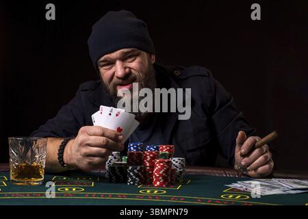 Bärtiger Mann in einem dunklen blauen Hut mit Zigarre und Glas am Poker Tisch sitzen und schreien auf Schwarz isoliert. Das Konzept des Glücksspiels Stockfoto