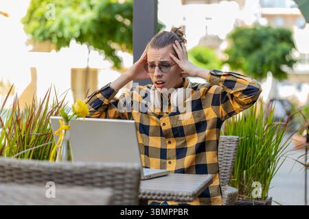 Gutaussehender junger Mann, der Laptop-Computer benutzt, um zu surfen, verliert überraschte plötzliche Lotterieergebnisse, schlechte Nachrichten, Verlust von Vermögen, scheitern. Guy sitzt draußen auf der Terrasse des städtischen Cafés. Lebensstil in der Stadt. Stockfoto