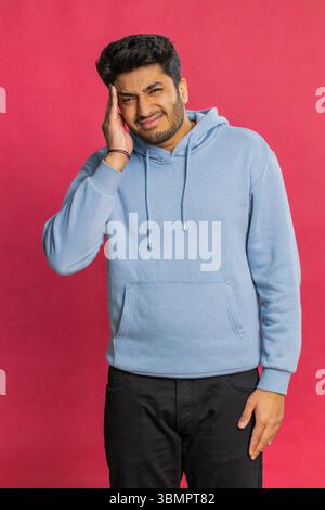 Unzufrieden erschöpfter indischer Hindu-Mann, der Schläfen reibt, um Kopfschmerzen zu heilen, leidet unter Spannung, Migräne, Stress, grimmigen Schmerzen, Bluthochdruck isoliert auf rotem Hintergrund Stockfoto